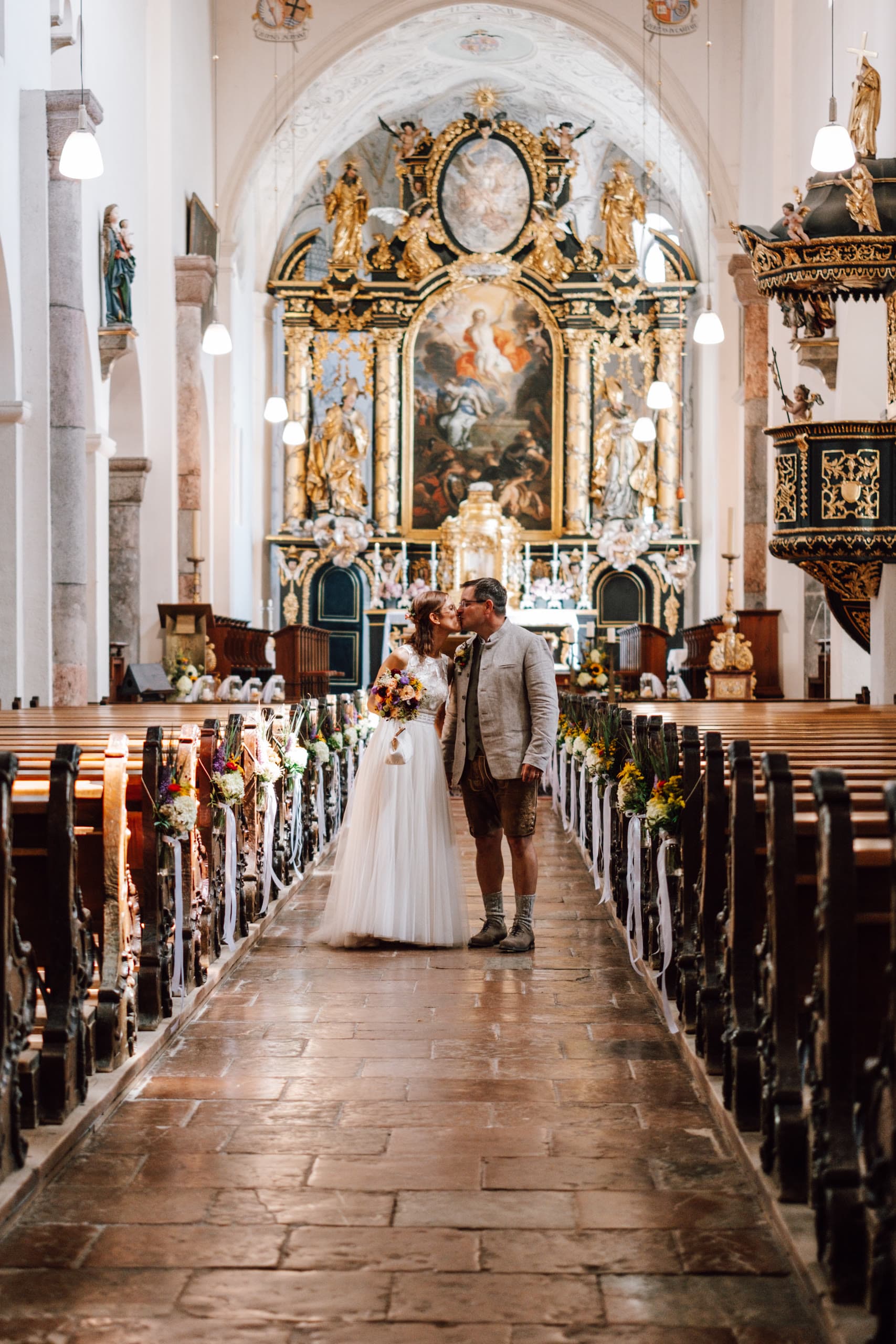 Bride and groom kiss in the aisle of a baroque church with a gilded altar behind