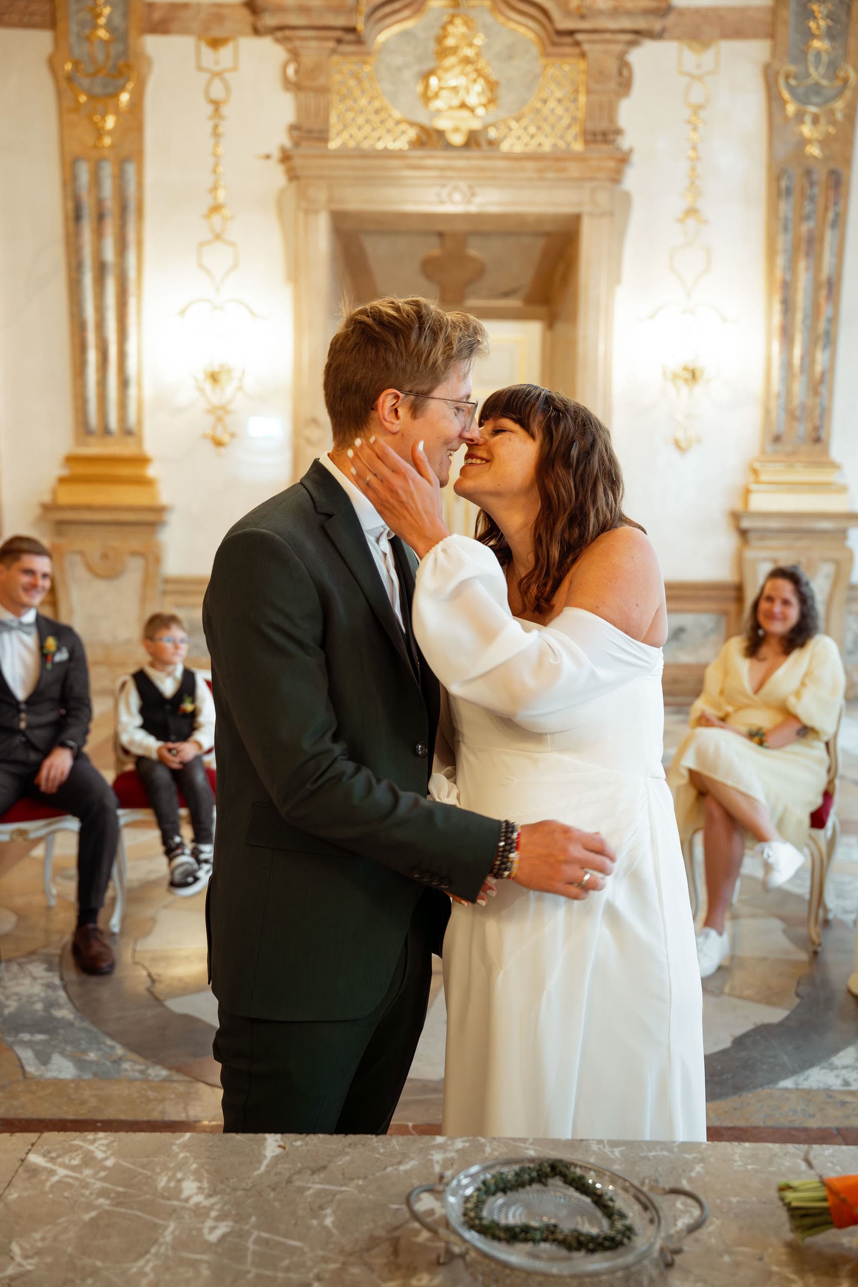 Bride and groom lean in to kiss after their civil ceremony in a gilded baroque hall, guests seated behind