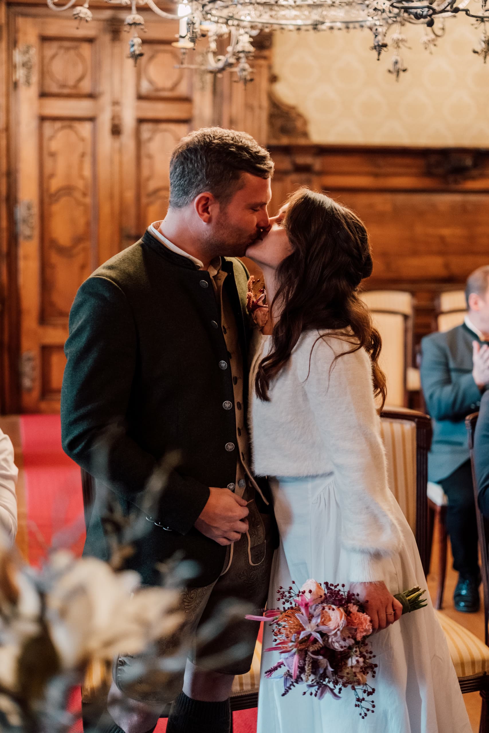 Groom in a green Tracht jacket kisses the bride in a wood-panelled room, guests seated behind