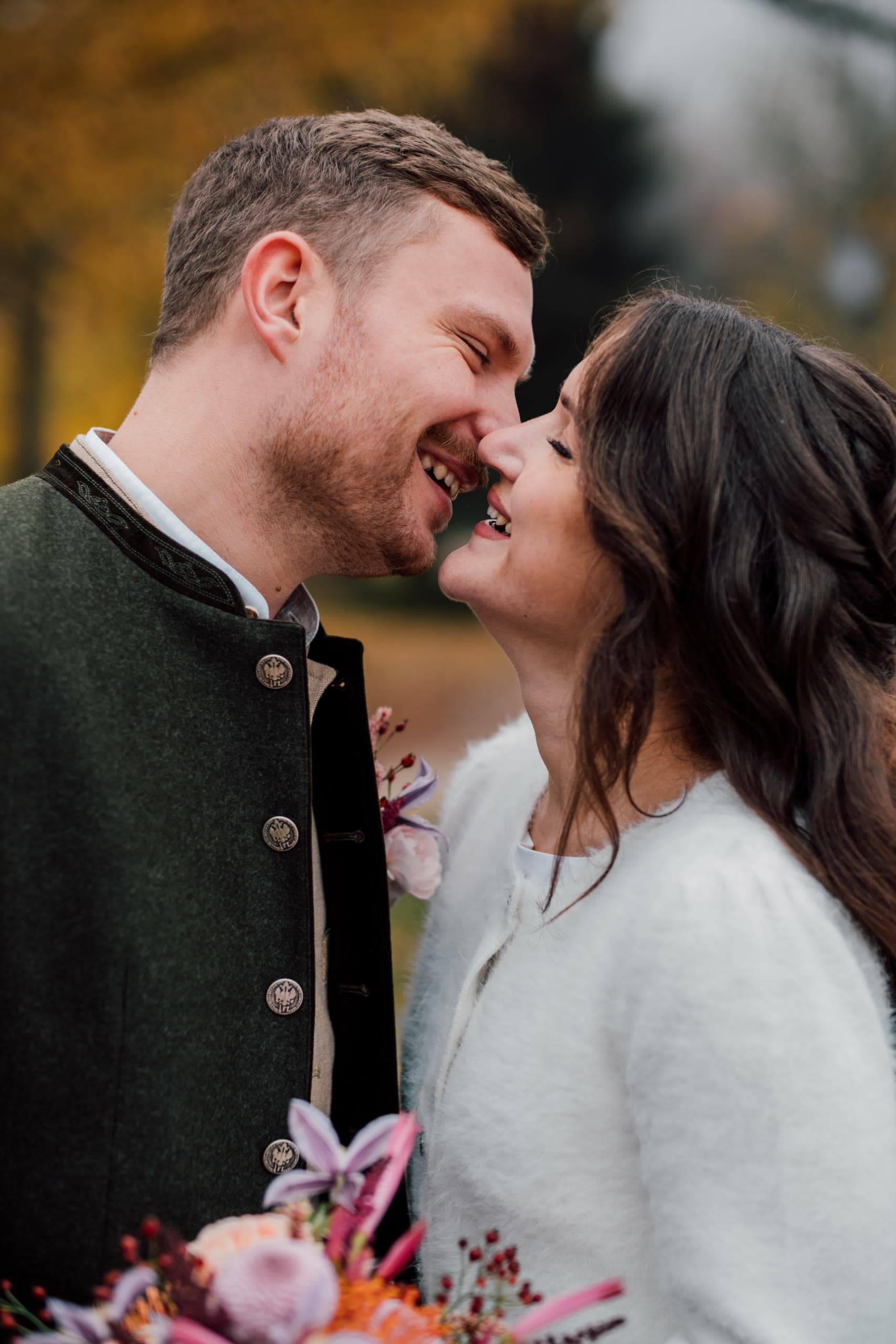 Groom in a green Tracht jacket and bride touch noses laughing, autumn foliage behind
