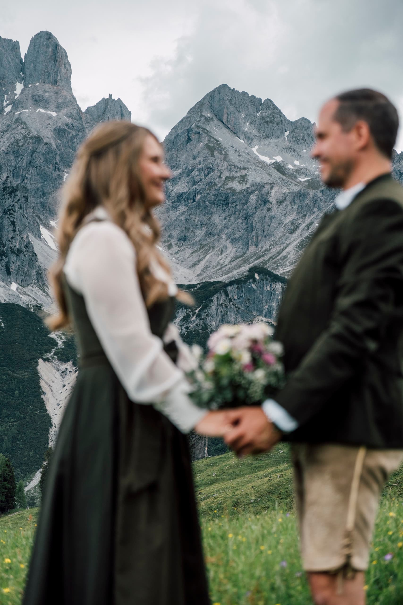 Bride in a dirndl and groom in Tracht hold hands facing each other, grey peaks blurred behind
