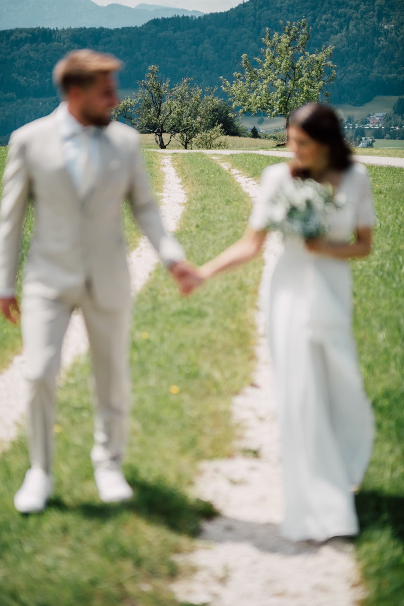 Bride and groom walk hand in hand along a dirt track with green fields behind