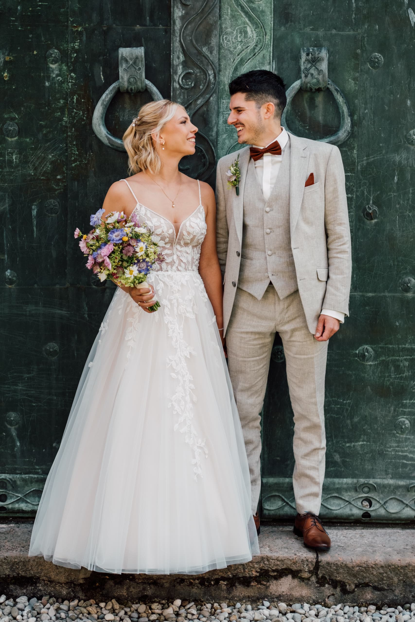 Bride and groom smile at each other before a weathered green double door with iron rings