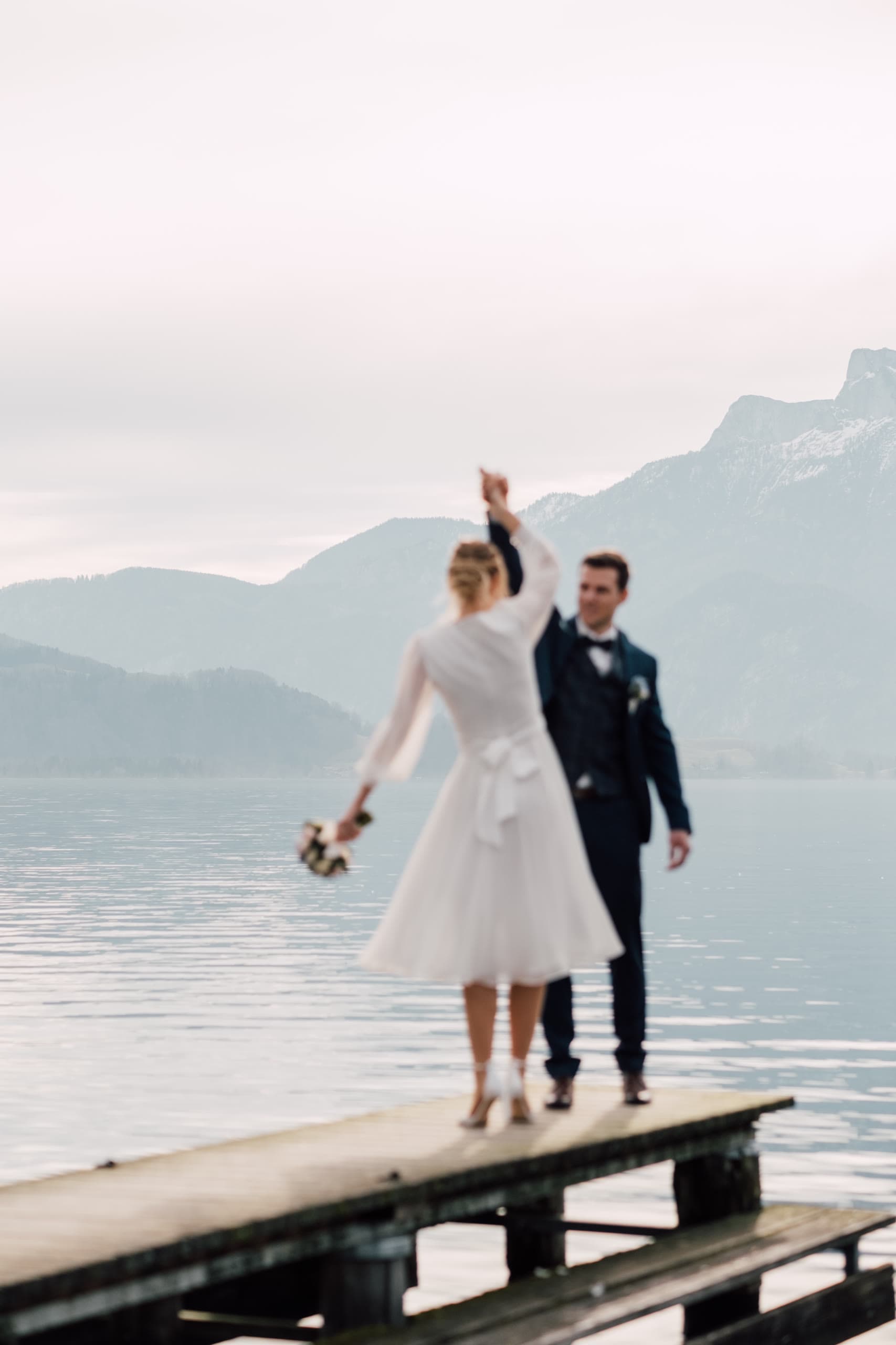 Bride walks along a lake dock with one arm raised, groom following in a dark suit