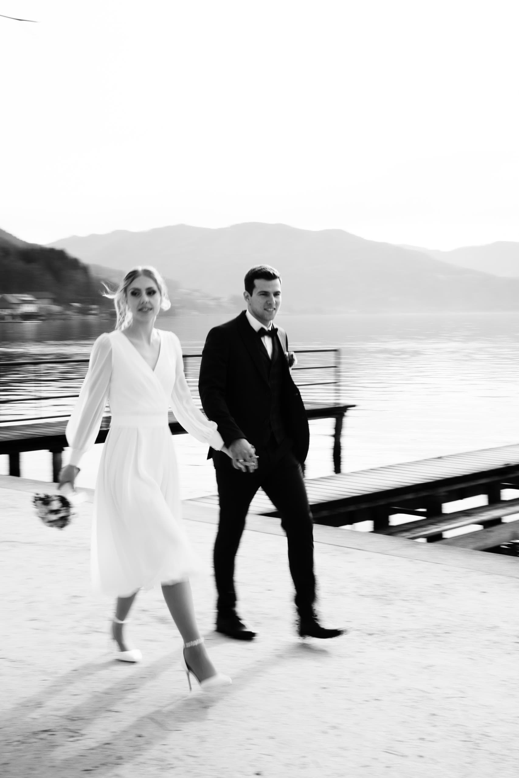 Motion-blurred black and white frame of a couple walking a lakeside dock, mountains behind