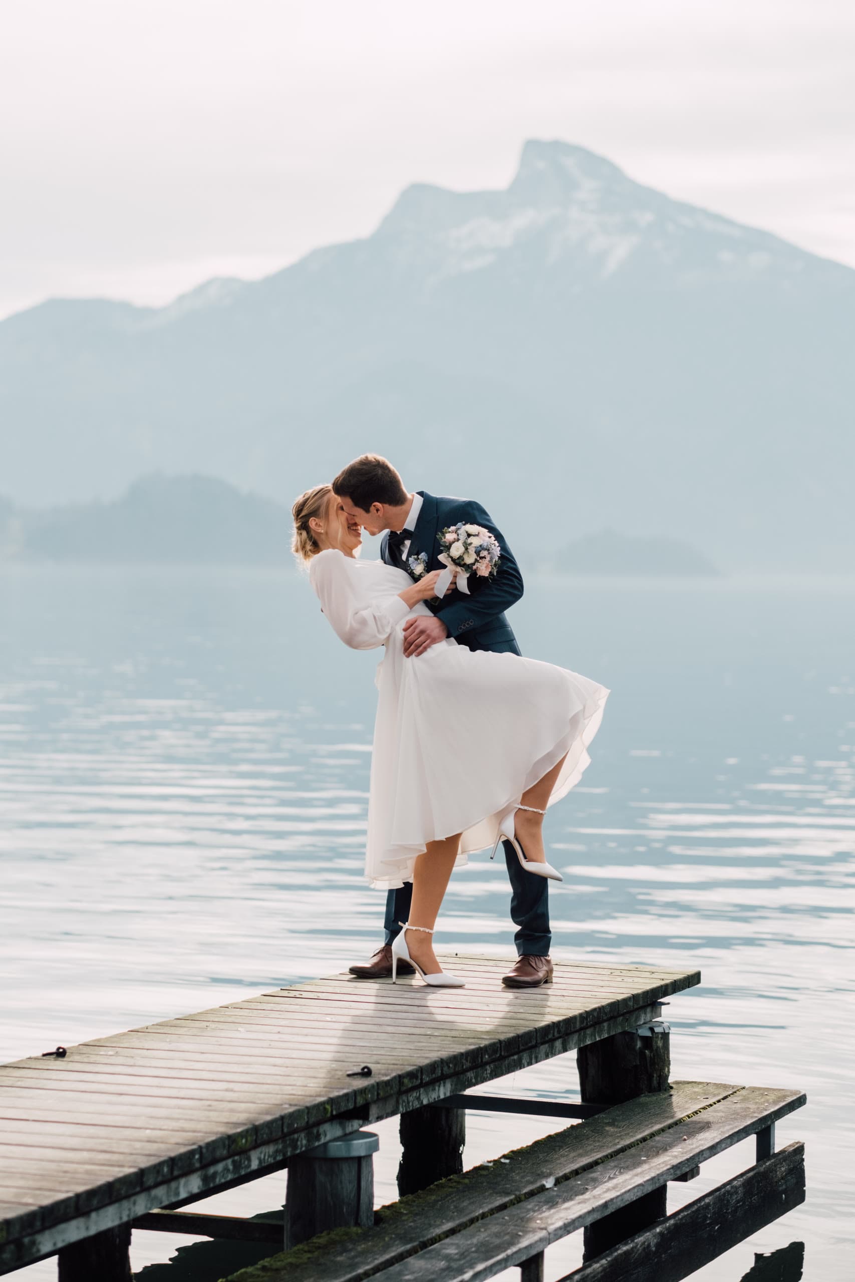 Groom dips the bride for a kiss on a lake dock, mountains behind them