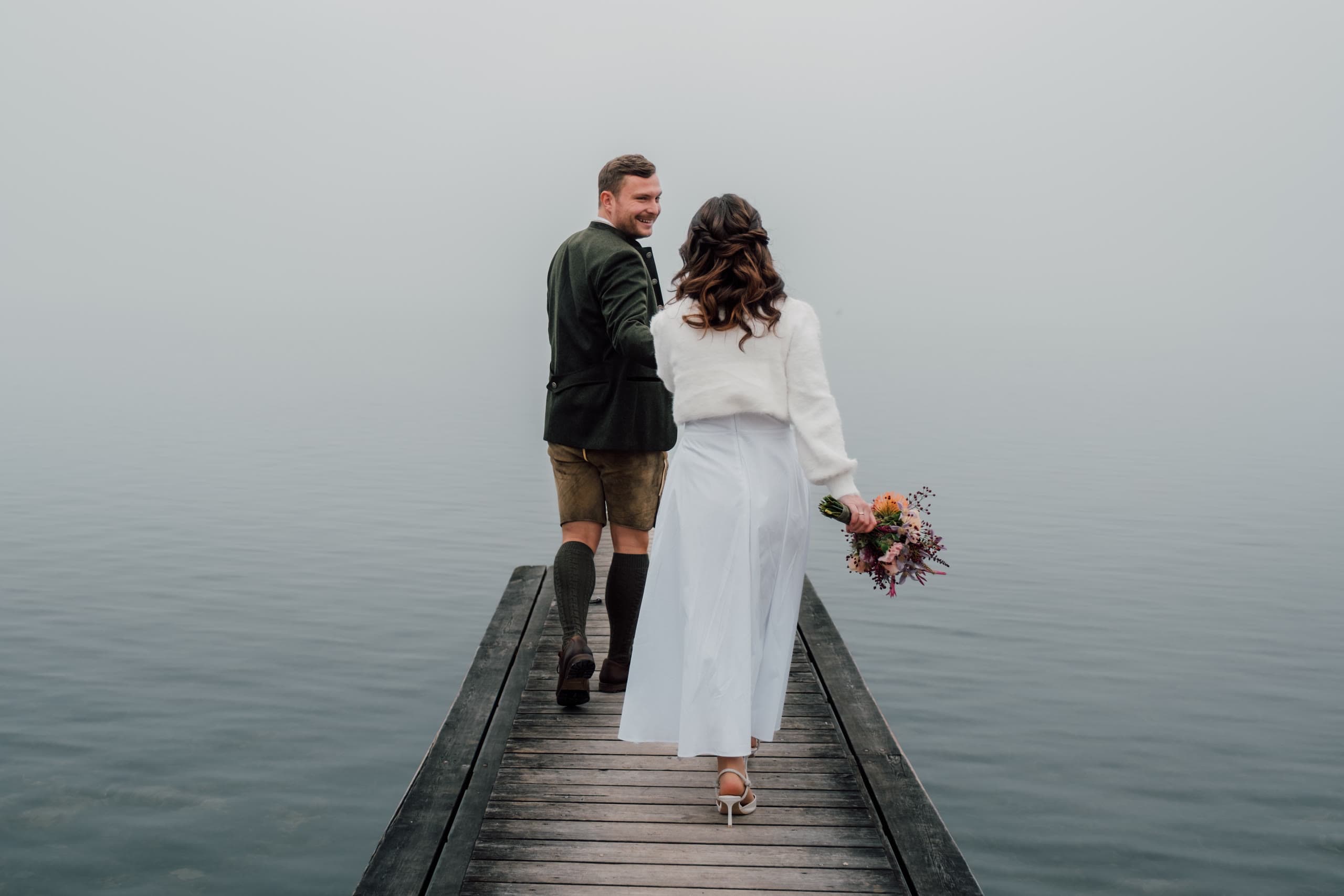 Couple walk away along a wooden dock into fog, bride in white, groom in Tracht