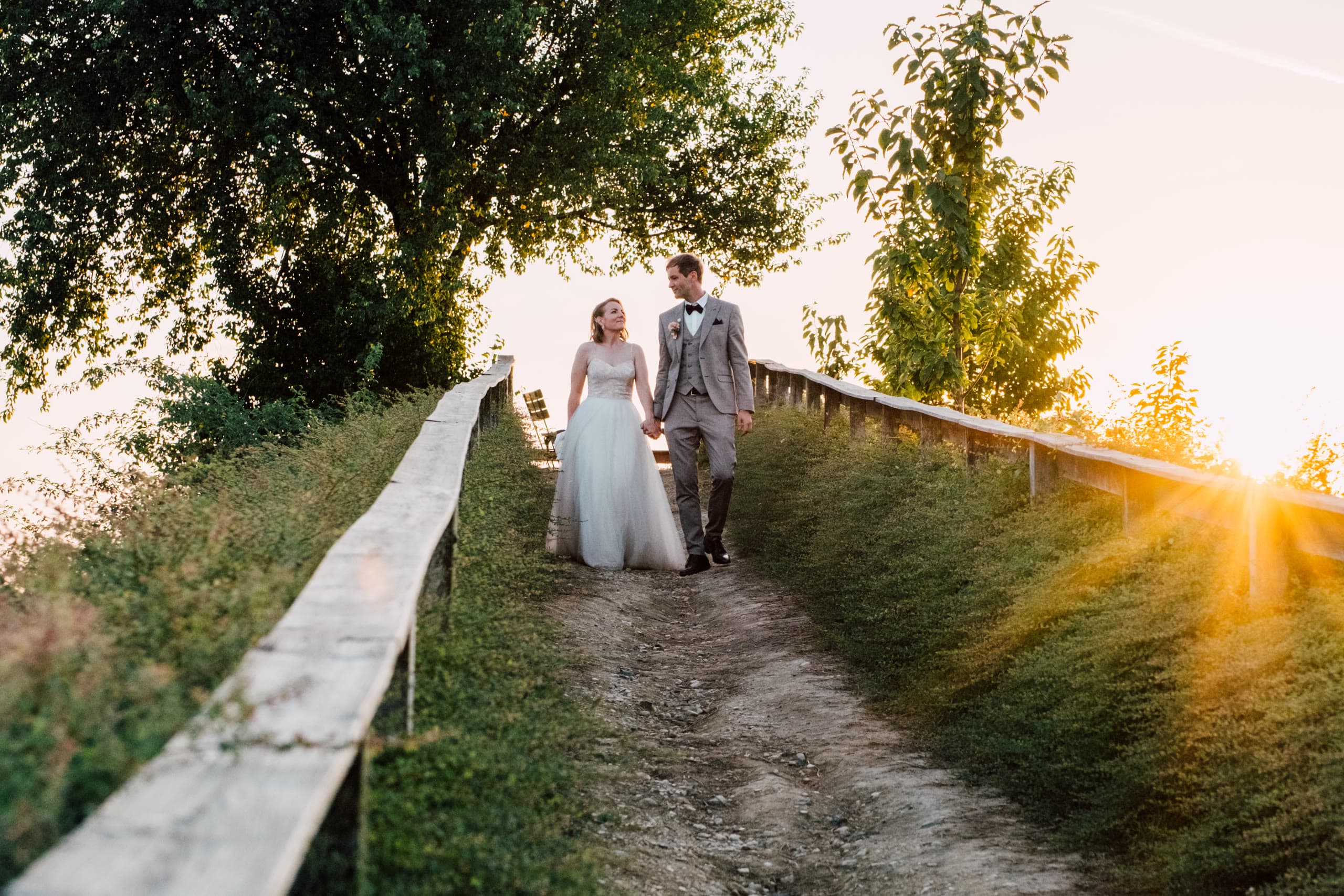 Couple walk hand in hand down a grassy path between wooden fences at sunset