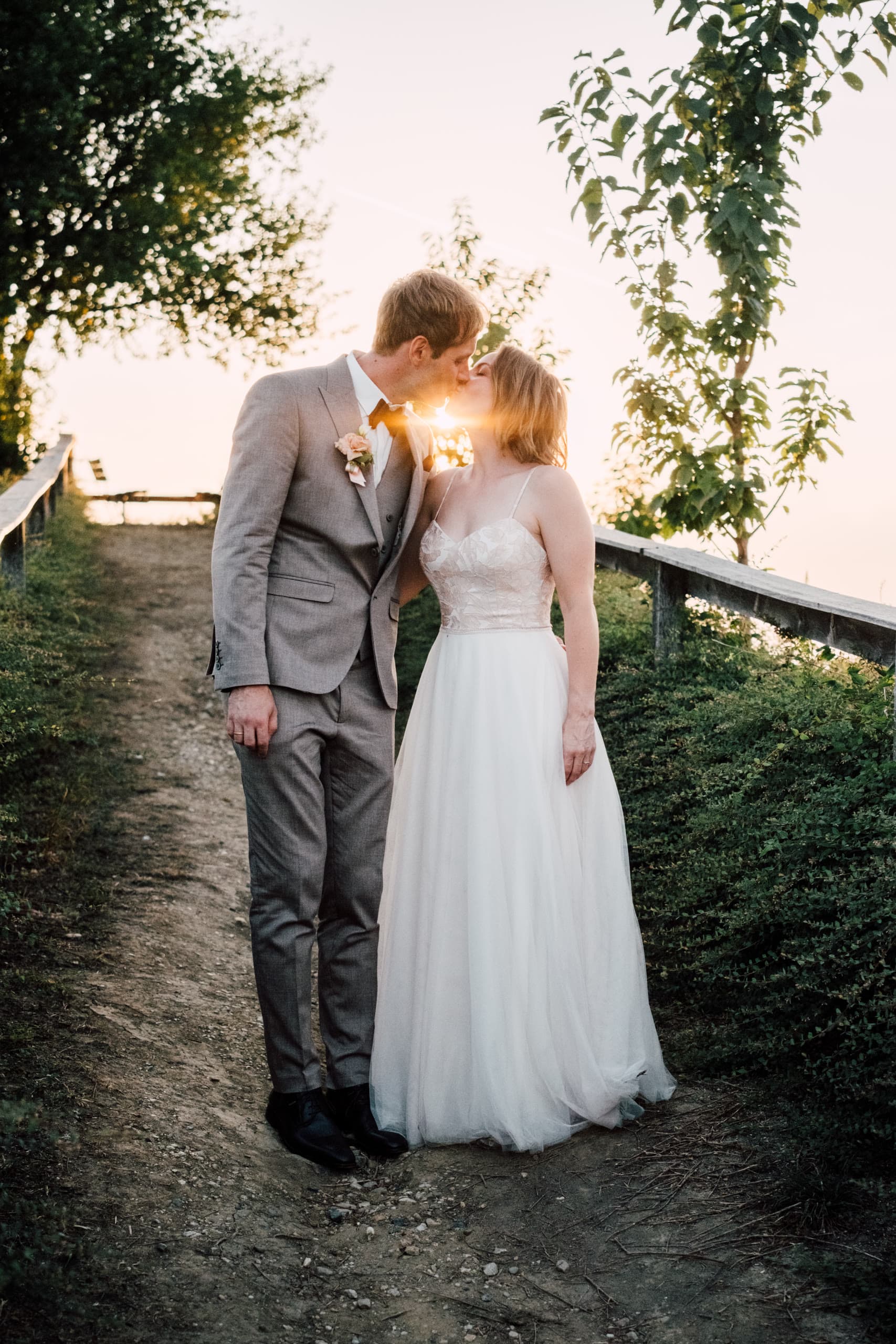 Couple kiss on a gravel hillside path lined by a wooden fence at sunset