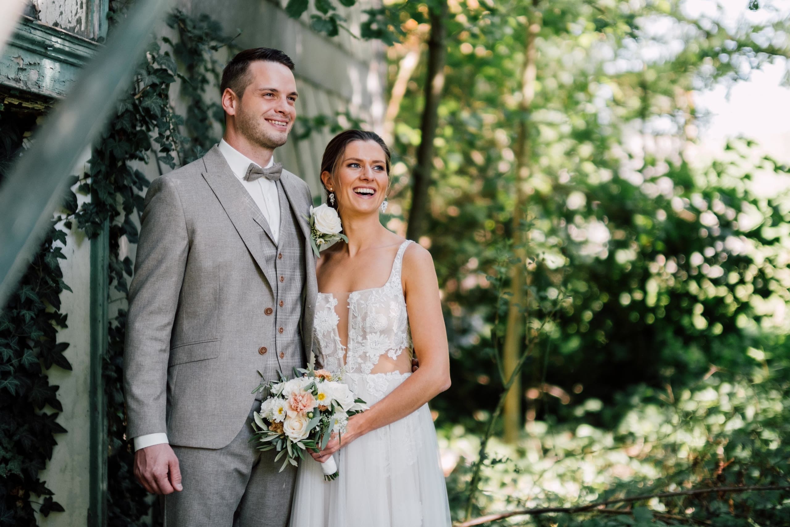 Bride and groom stand smiling beside an ivy-covered wall and a painted iron stair rail