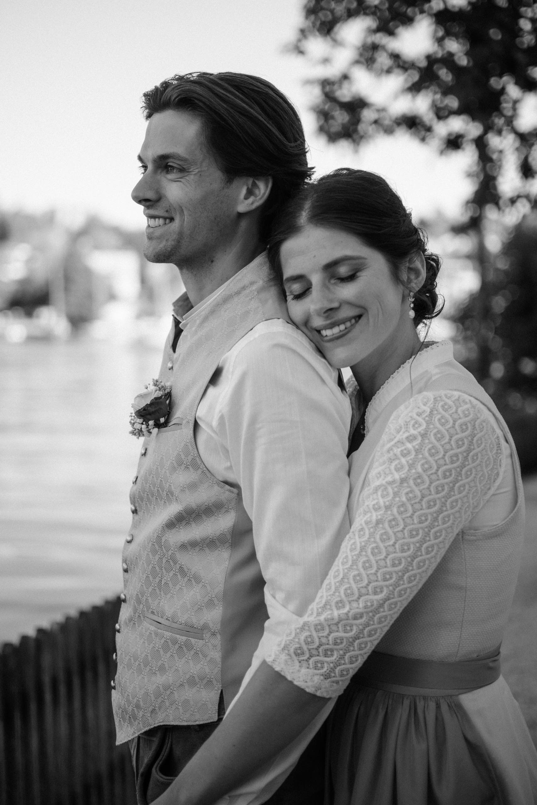 Groom in a waistcoat and bride embrace by a lake, her head resting on his shoulder
