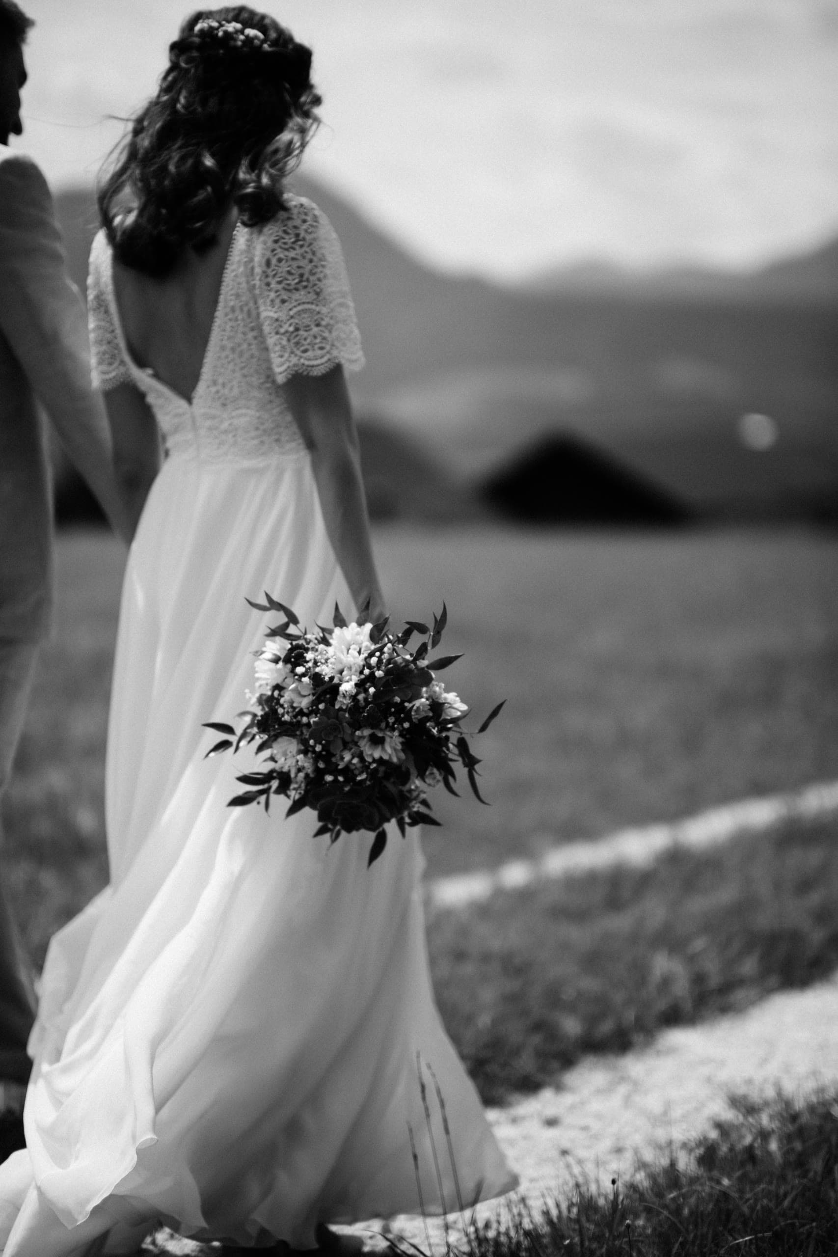 Bride walks away across a meadow in black and white, holding a bouquet and the groom's hand