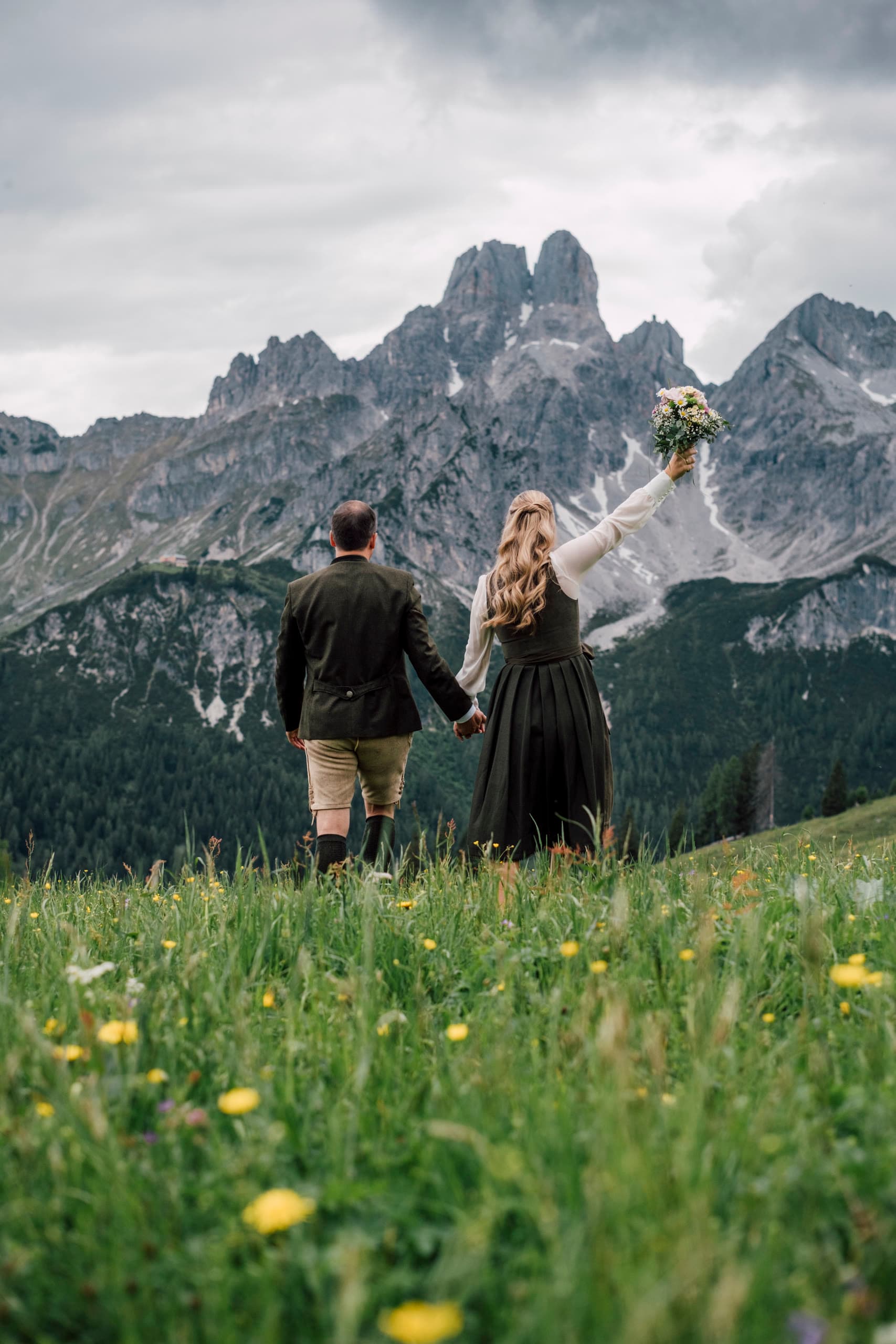 Couple in Tracht and dirndl walk away across a meadow toward grey peaks with snow patches