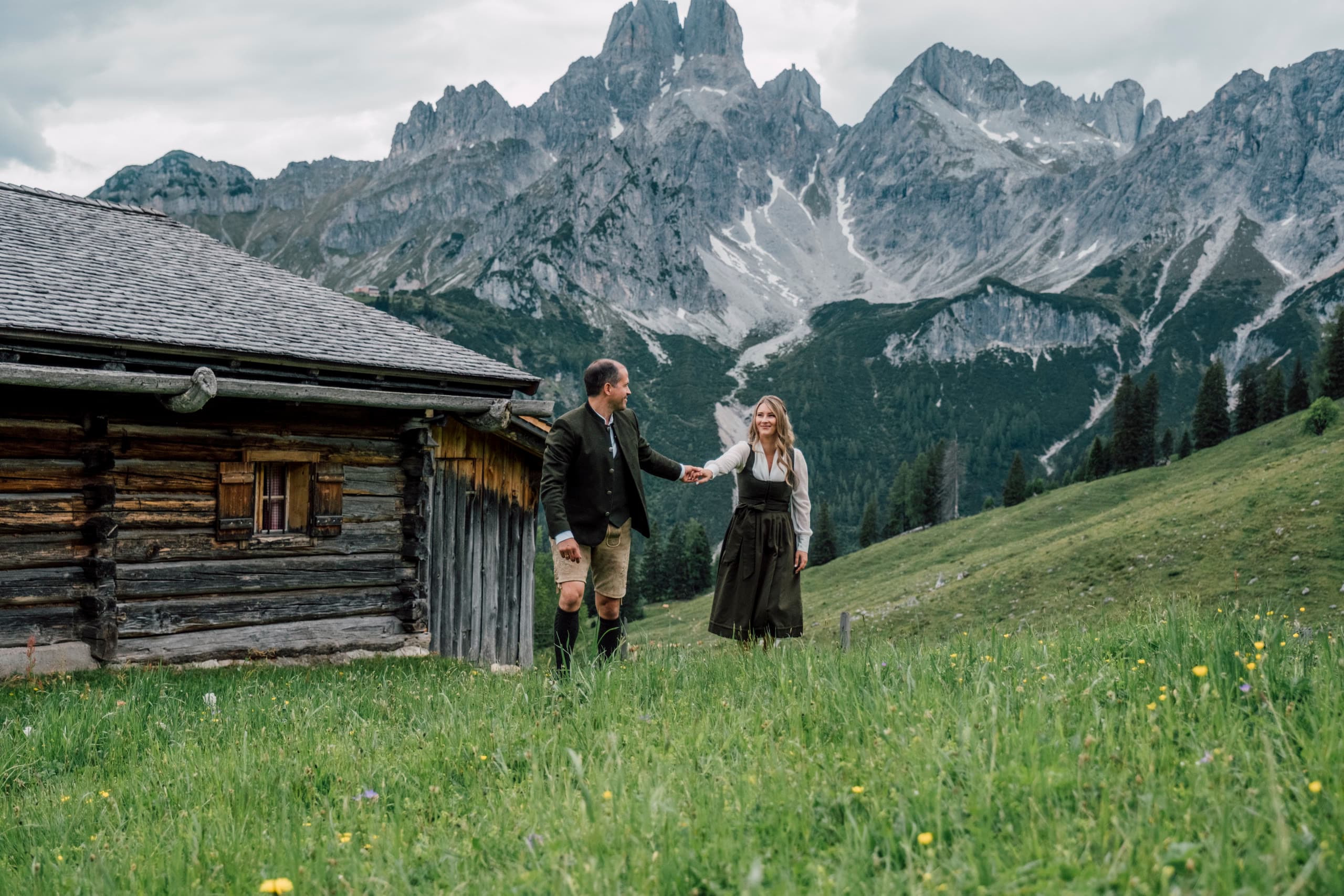 Couple in Tracht walk hand in hand across a meadow before a wooden hut and grey peaks
