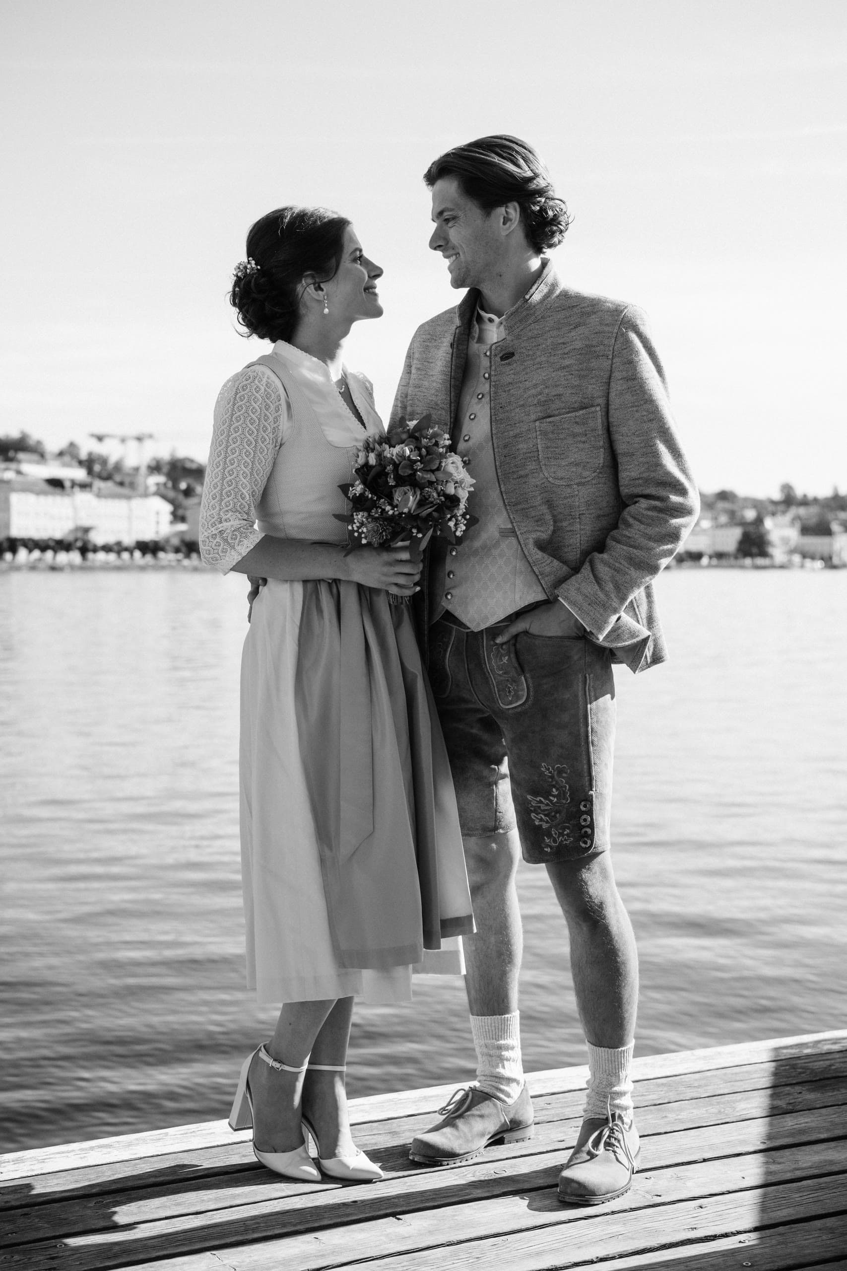 Couple in Tracht face each other on a wooden pier, water and a town behind, black and white