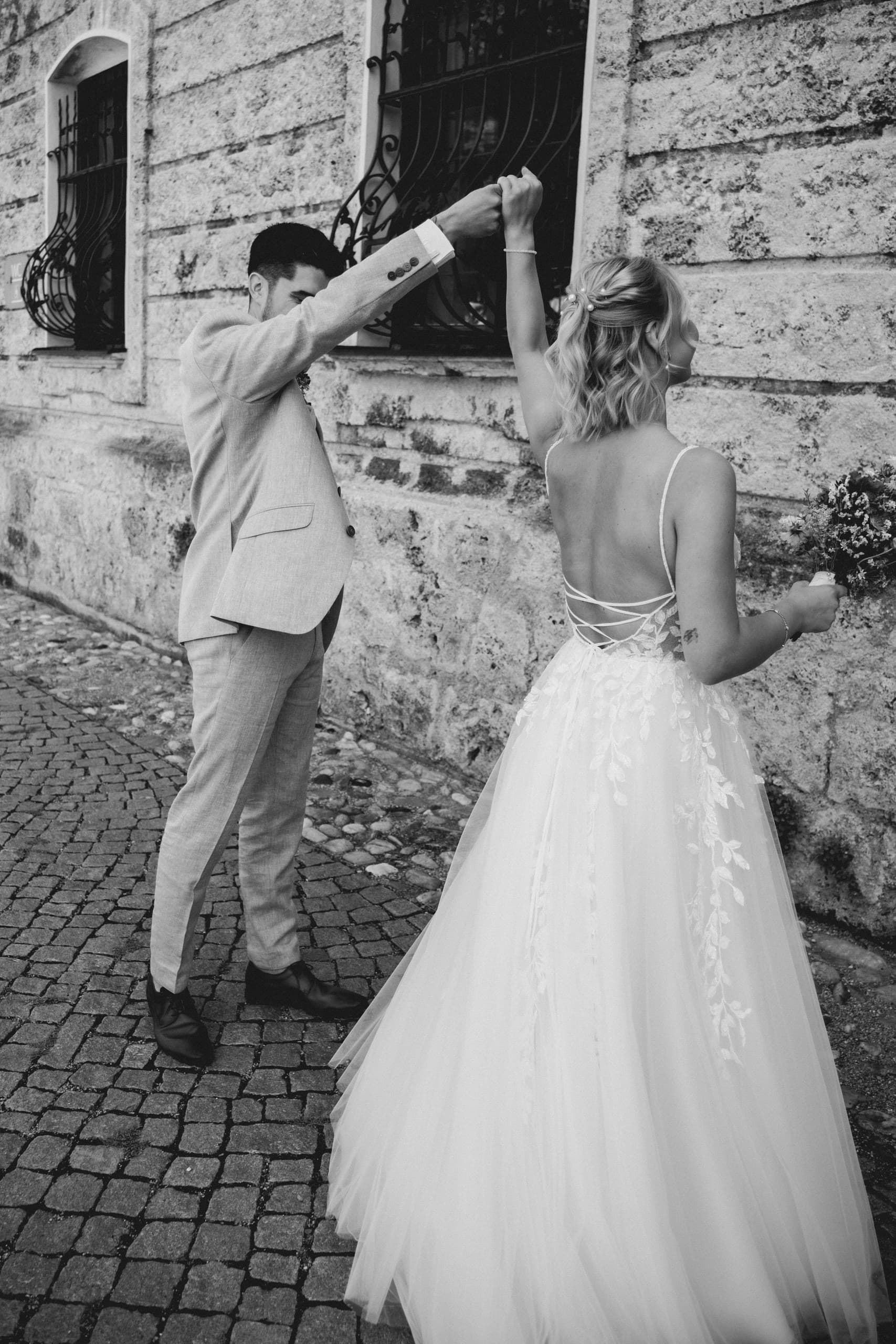 Couple dance with joined hands raised beside an old stone building, in black and white