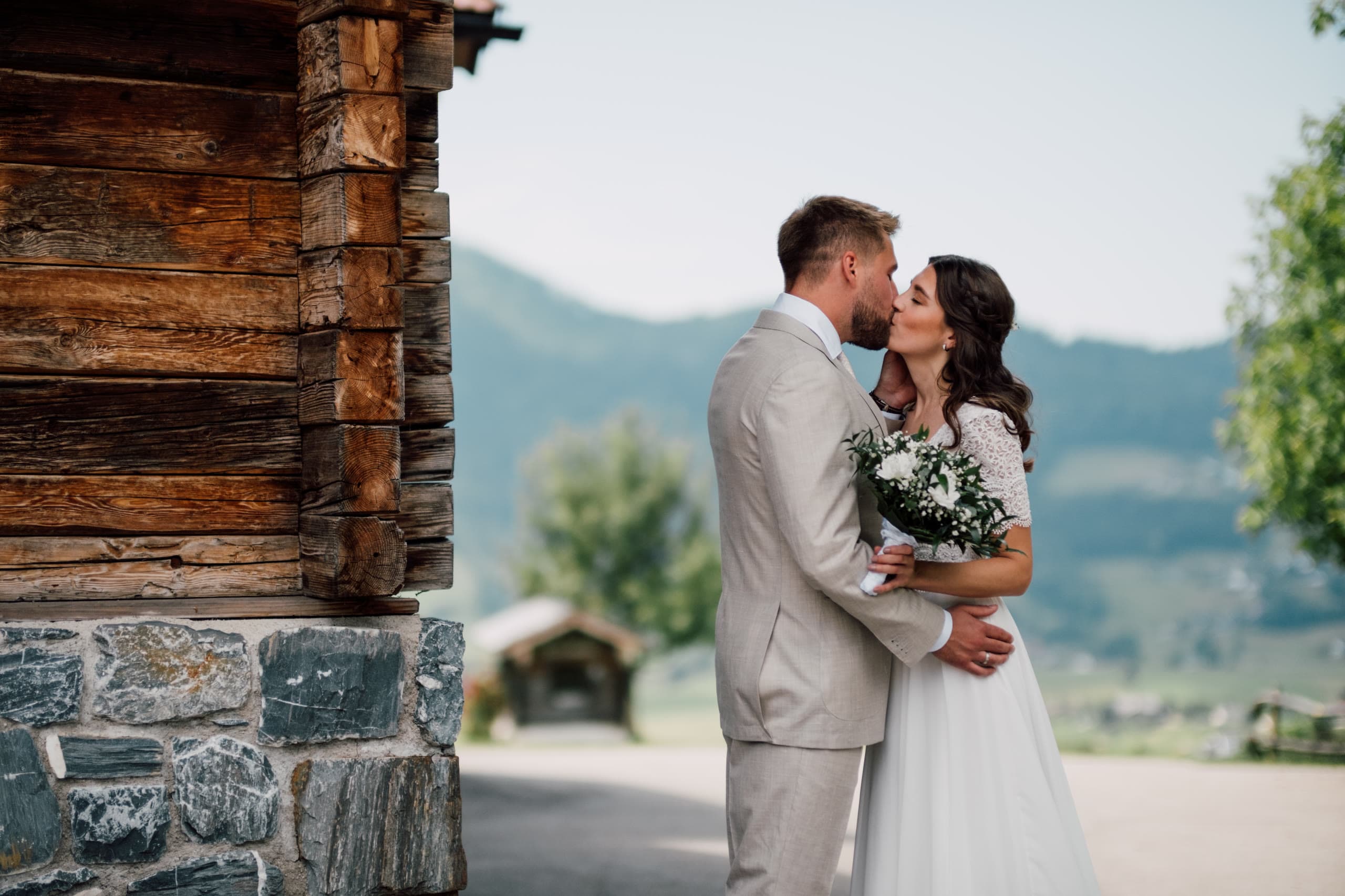 Couple kiss beside a timber and stone building with meadow and hazy hills behind