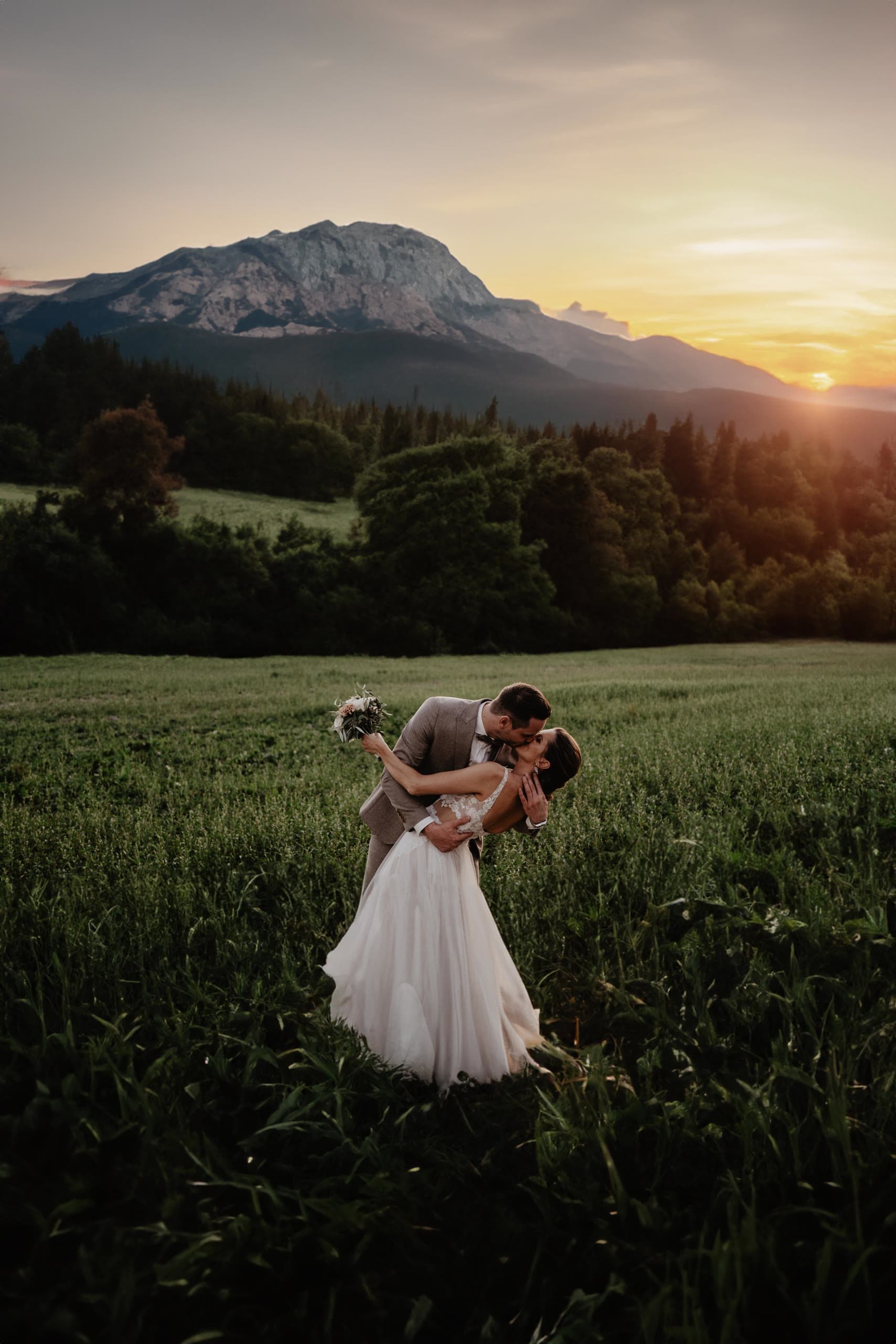 Groom dips the bride for a kiss in a tall meadow, a mountain peak and sunset behind them