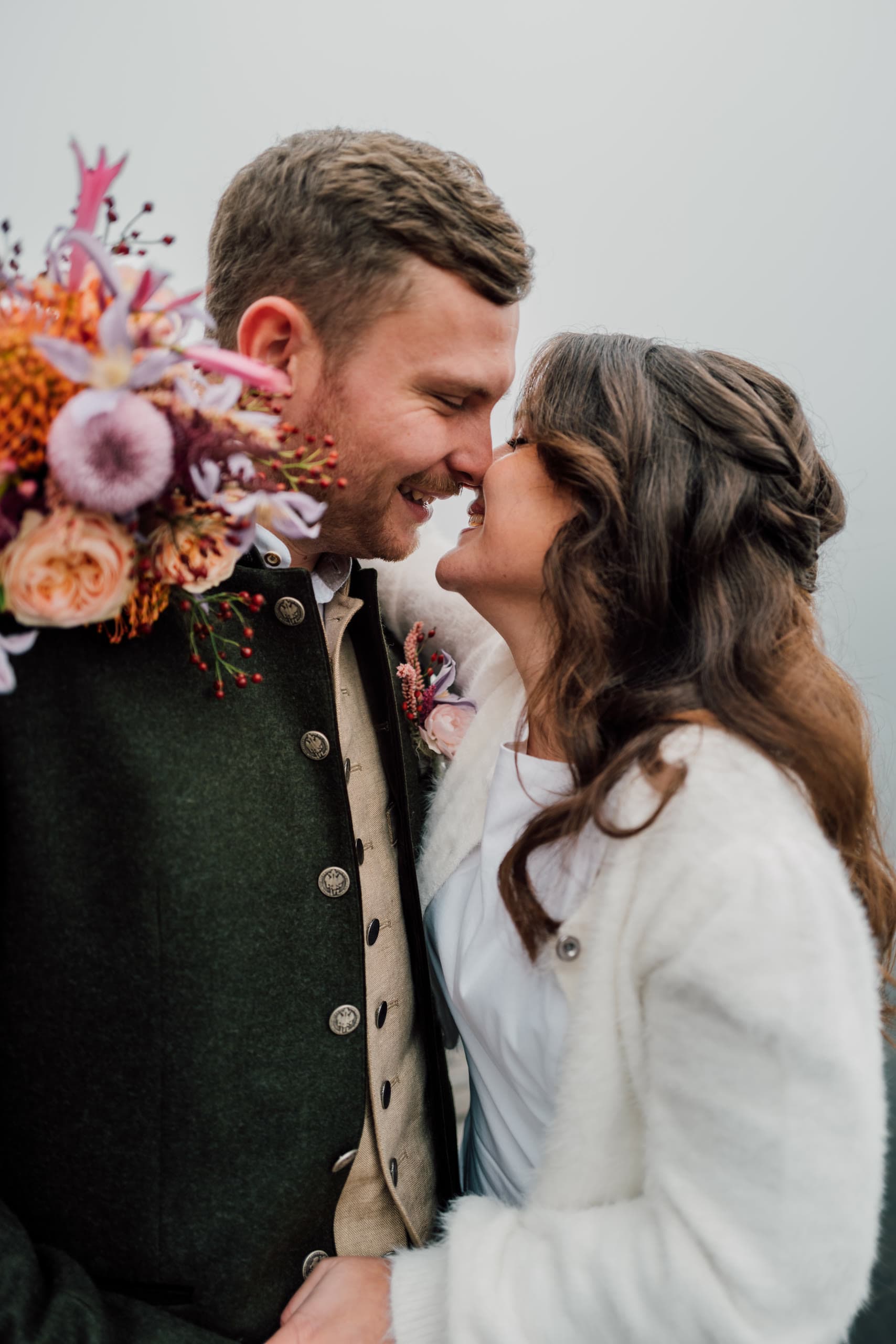 Couple in Tracht smile nose to nose with a bright bouquet held between them