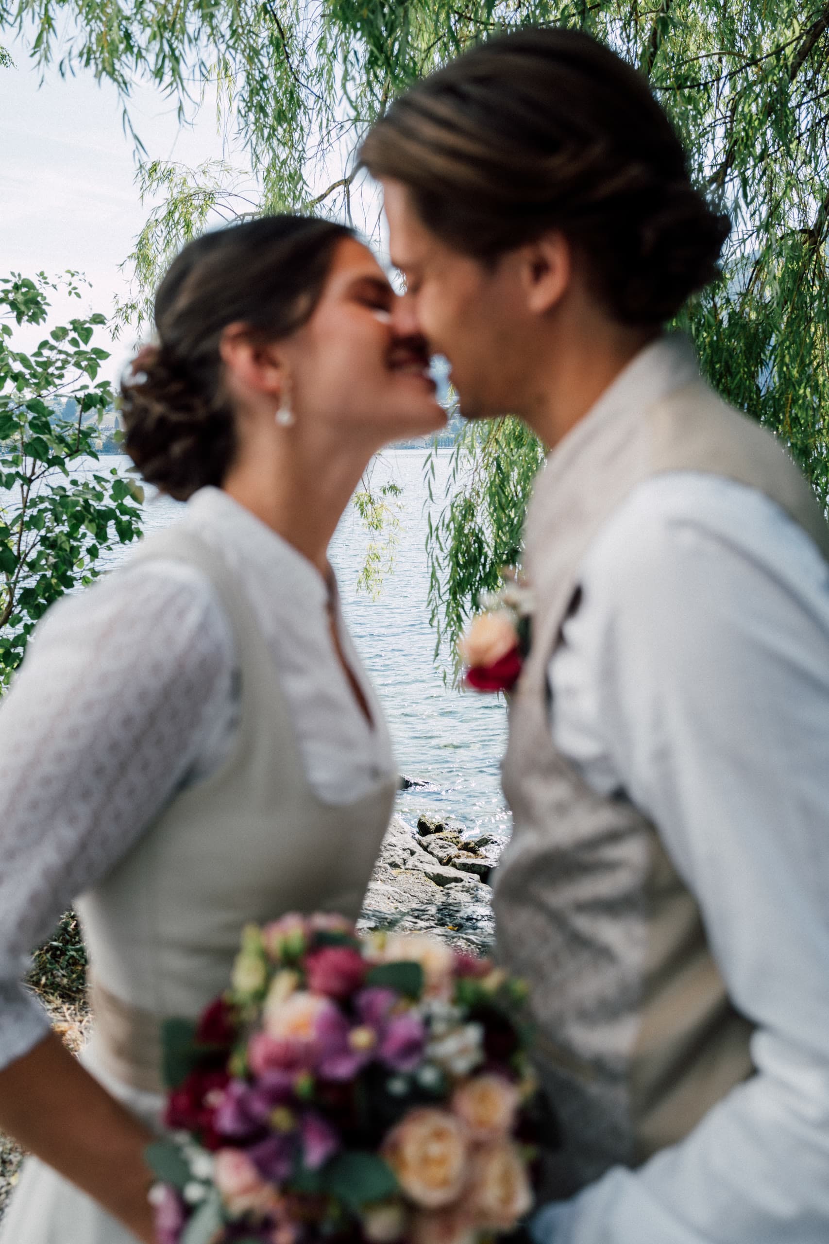 Bride and groom kiss under willow branches beside a lake, bride holding a bouquet