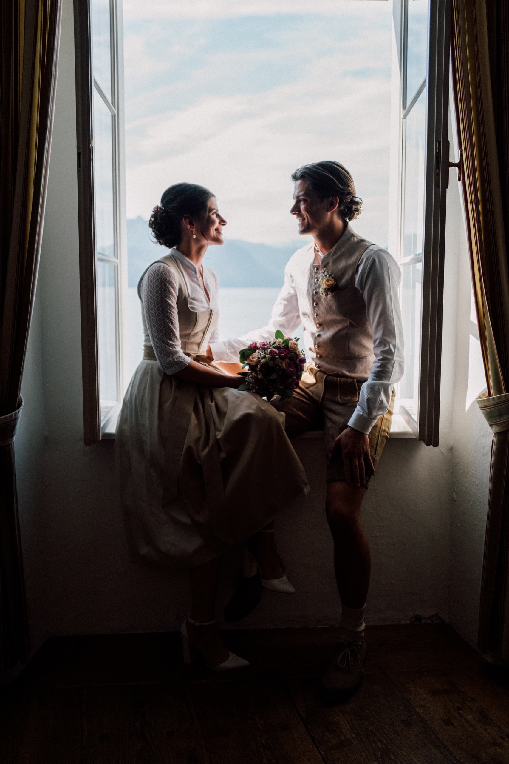 Couple in Austrian Tracht sit together in a window nook silhouetted against a lake
