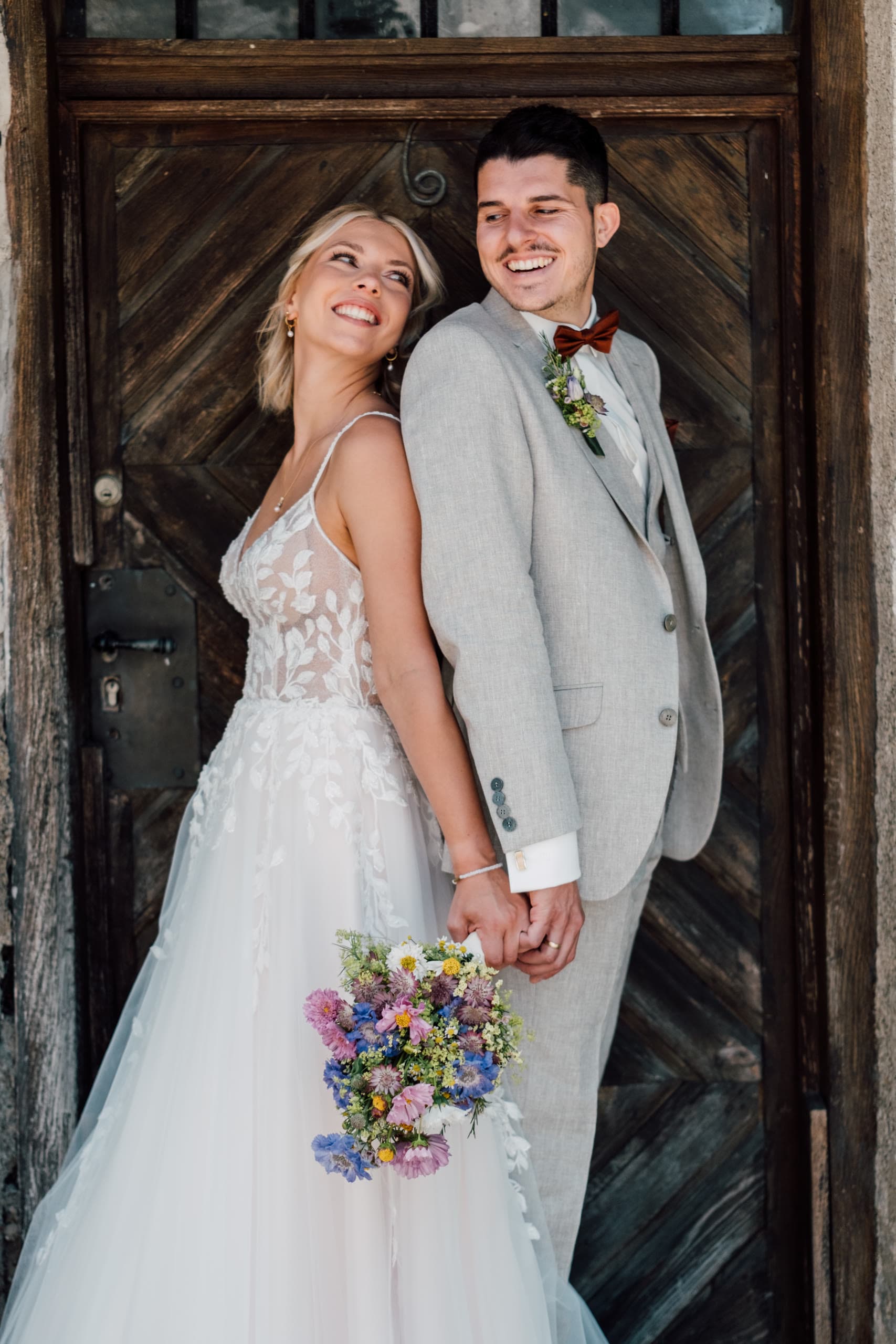 Bride and groom stand smiling before a weathered wooden door, holding hands