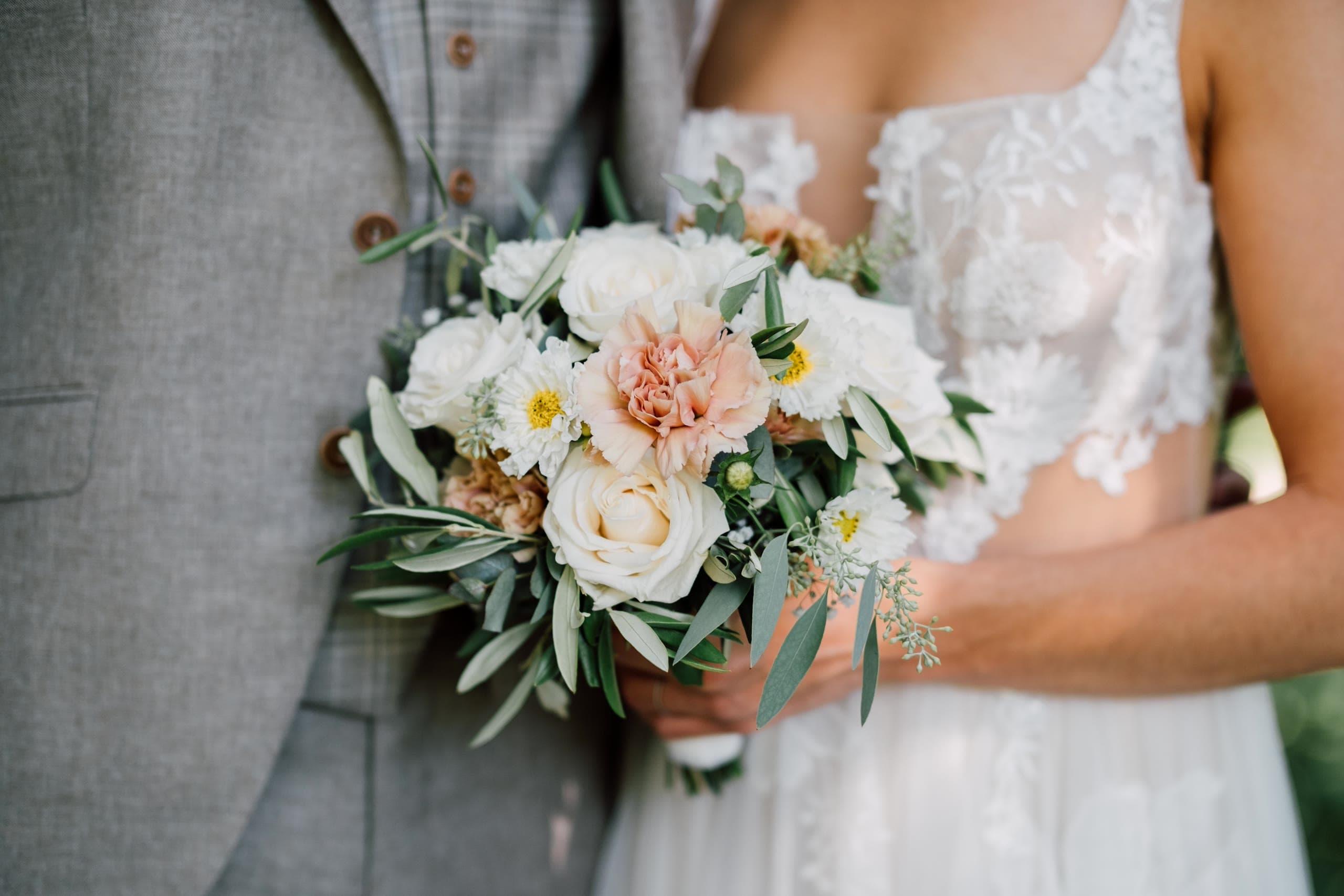 Bride holds a bouquet of white roses, peach carnations and olive leaves against the groom's grey suit
