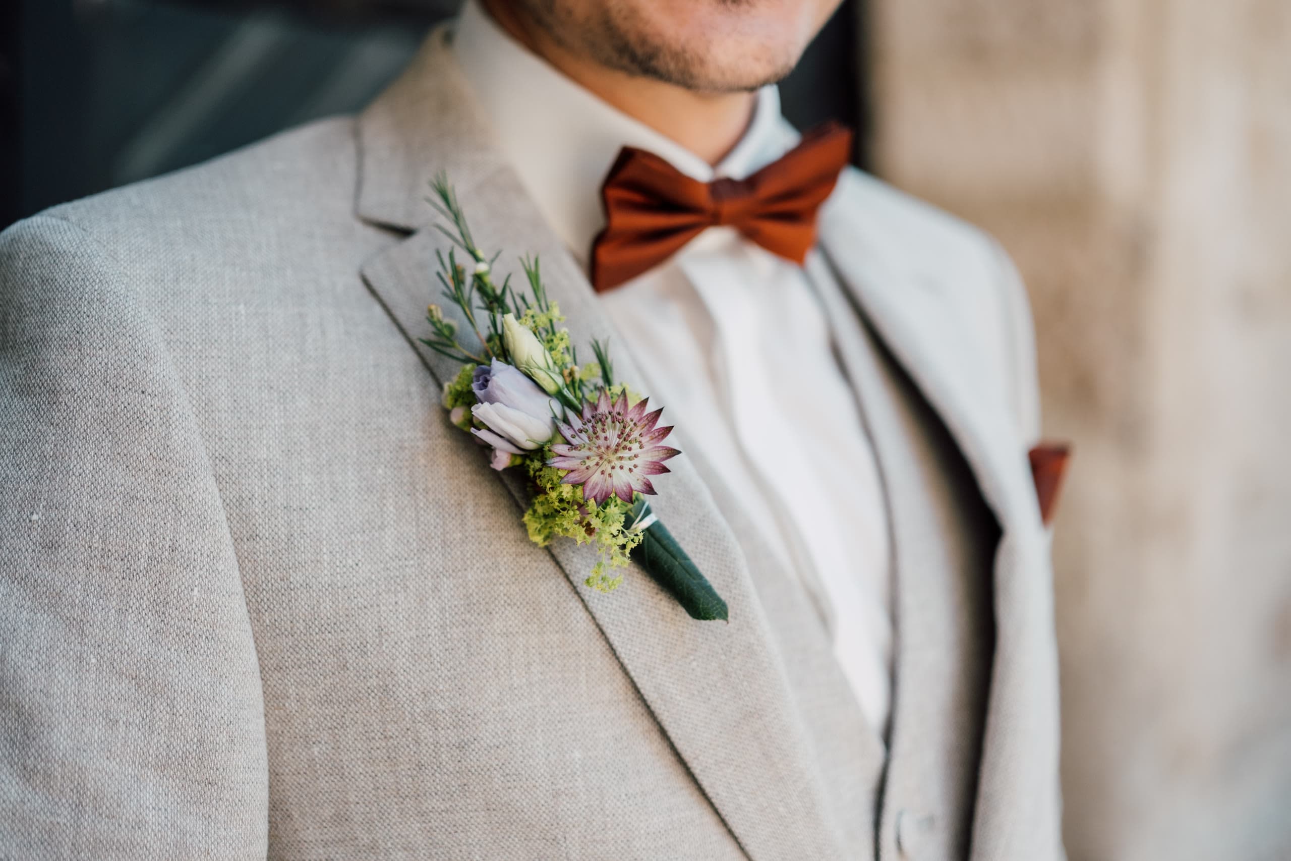 Close-up of a groom's boutonniere with purple thistle and sage against a beige jacket