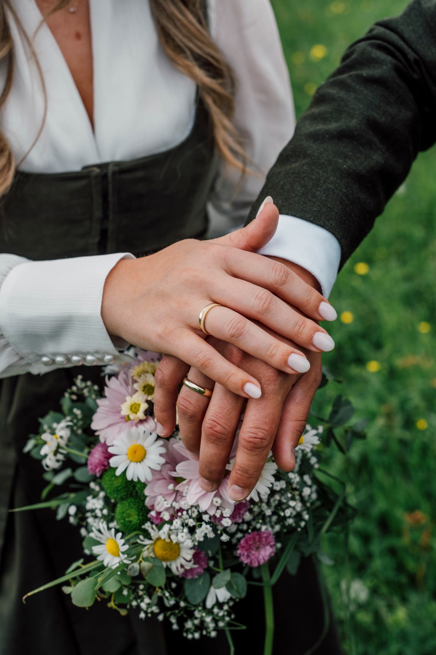 Bride and groom's hands rest on a wildflower bouquet, both wedding rings visible