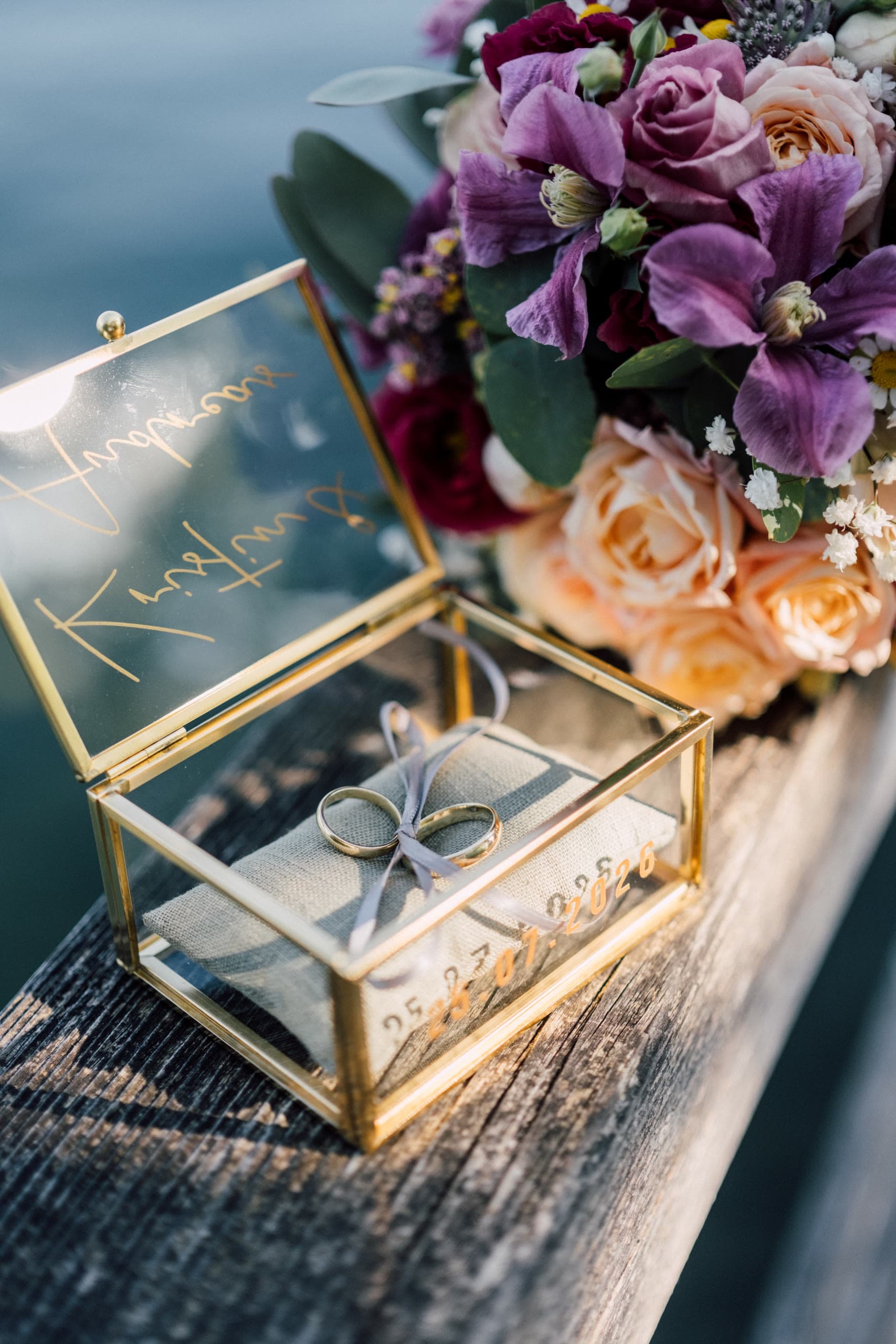 Two gold wedding rings in an open glass box beside a bouquet on a dock