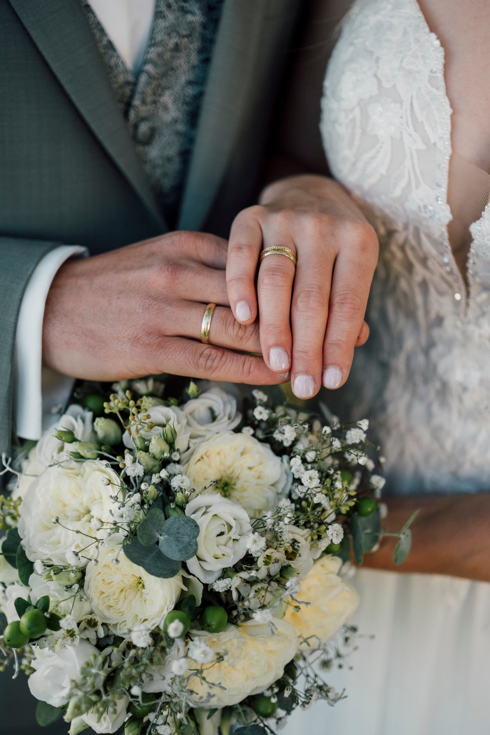 Bride and groom's ringed hands rest together above a bouquet of white roses