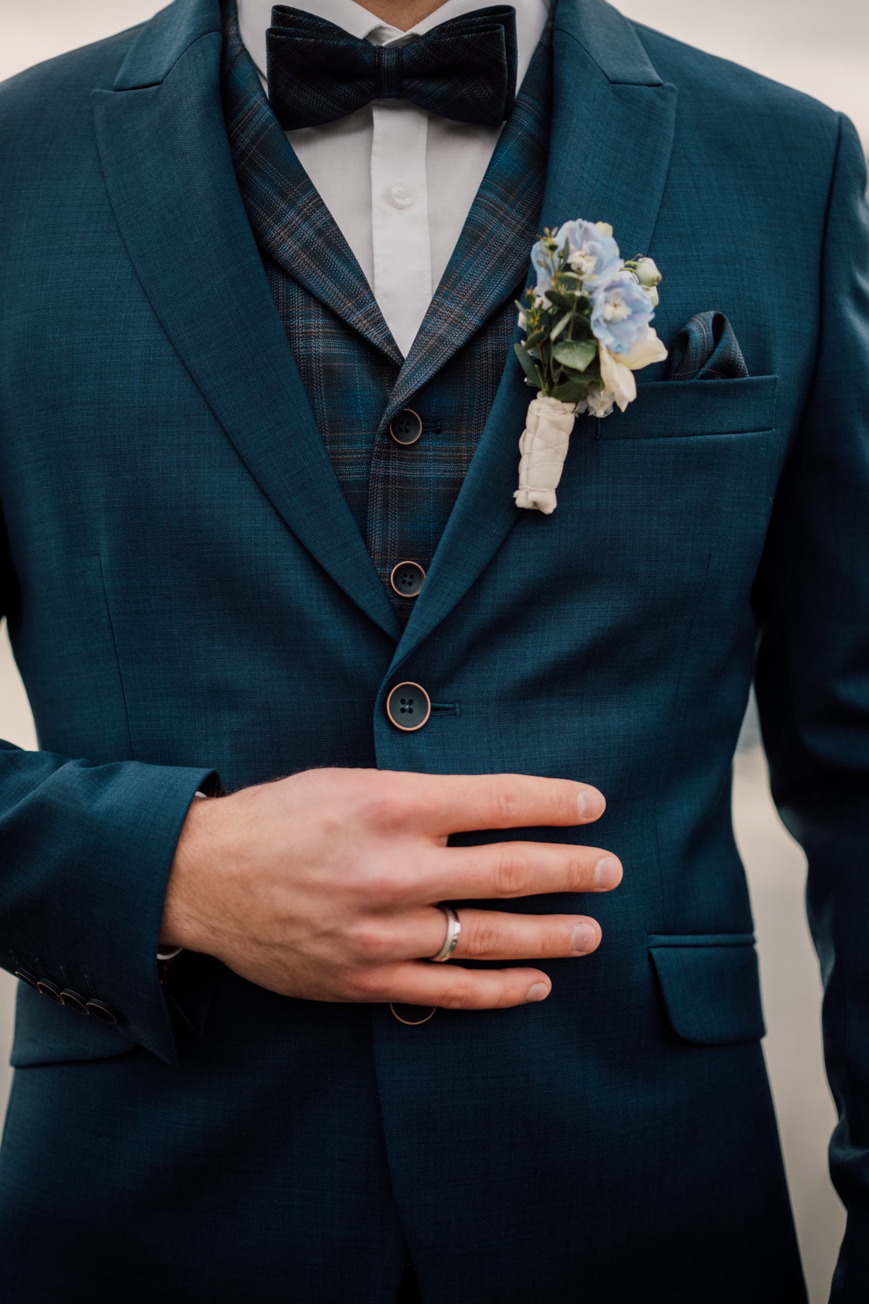 Close-up of a groom's teal suit jacket, hand on chest showing a wedding ring and boutonniere