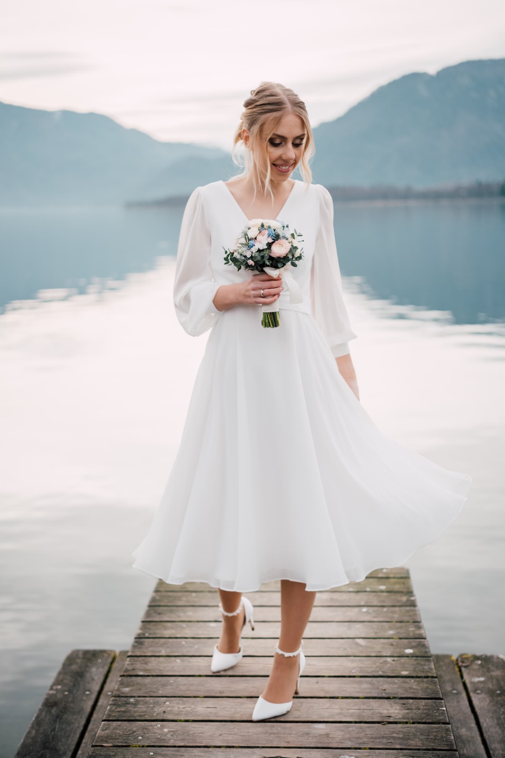 Bride in a tea-length dress walks a wooden dock over a lake, mountains beyond