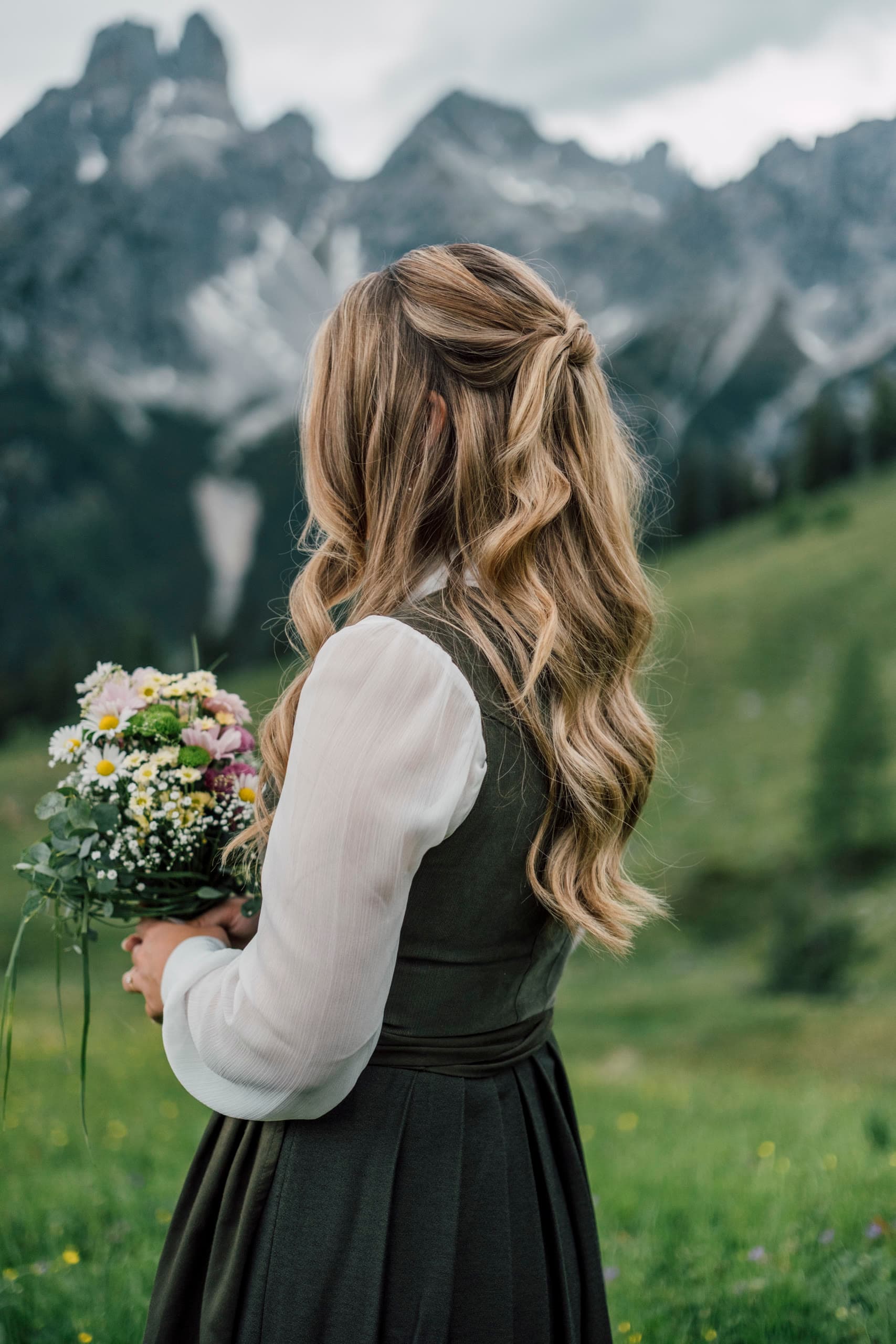 Woman in a traditional Tracht dress seen from behind, holding wildflowers, facing grey peaks