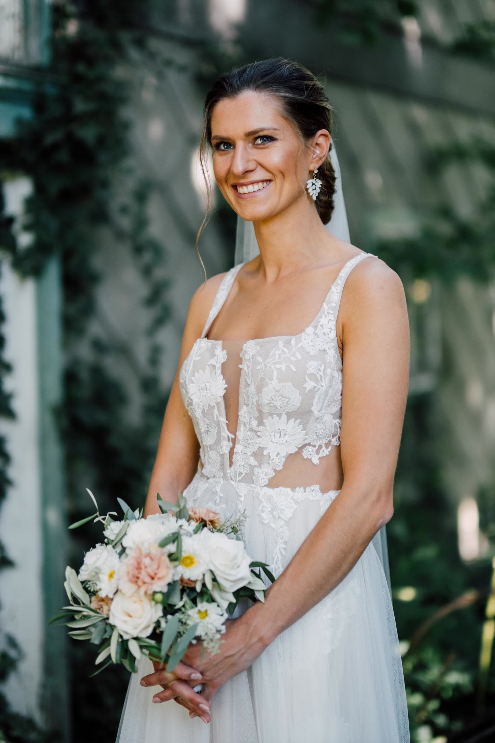 Bride in a floral lace dress and veil smiles at the camera, holding a white and peach bouquet