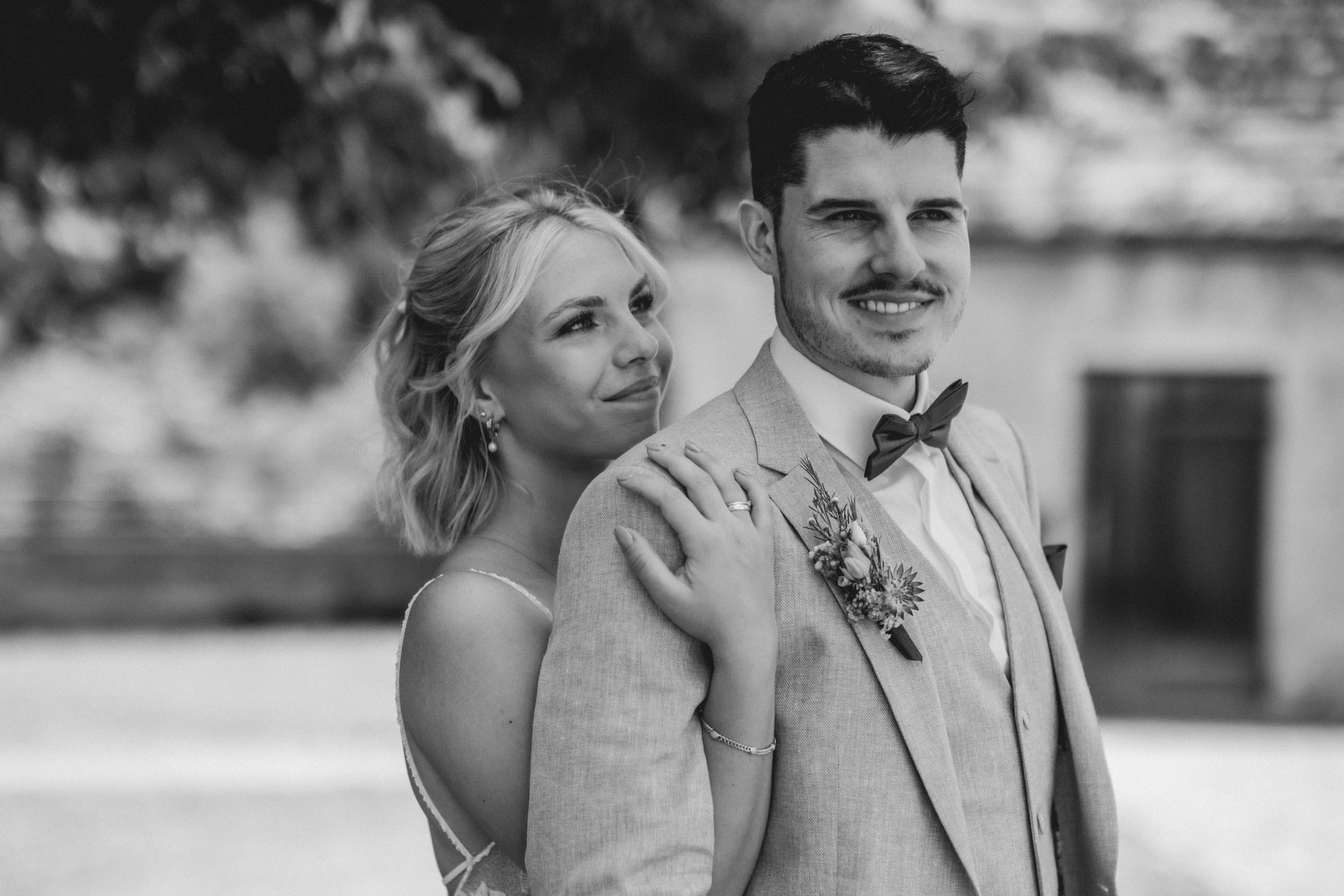 Bride rests her hand on the groom's shoulder, both smiling outdoors, in black and white