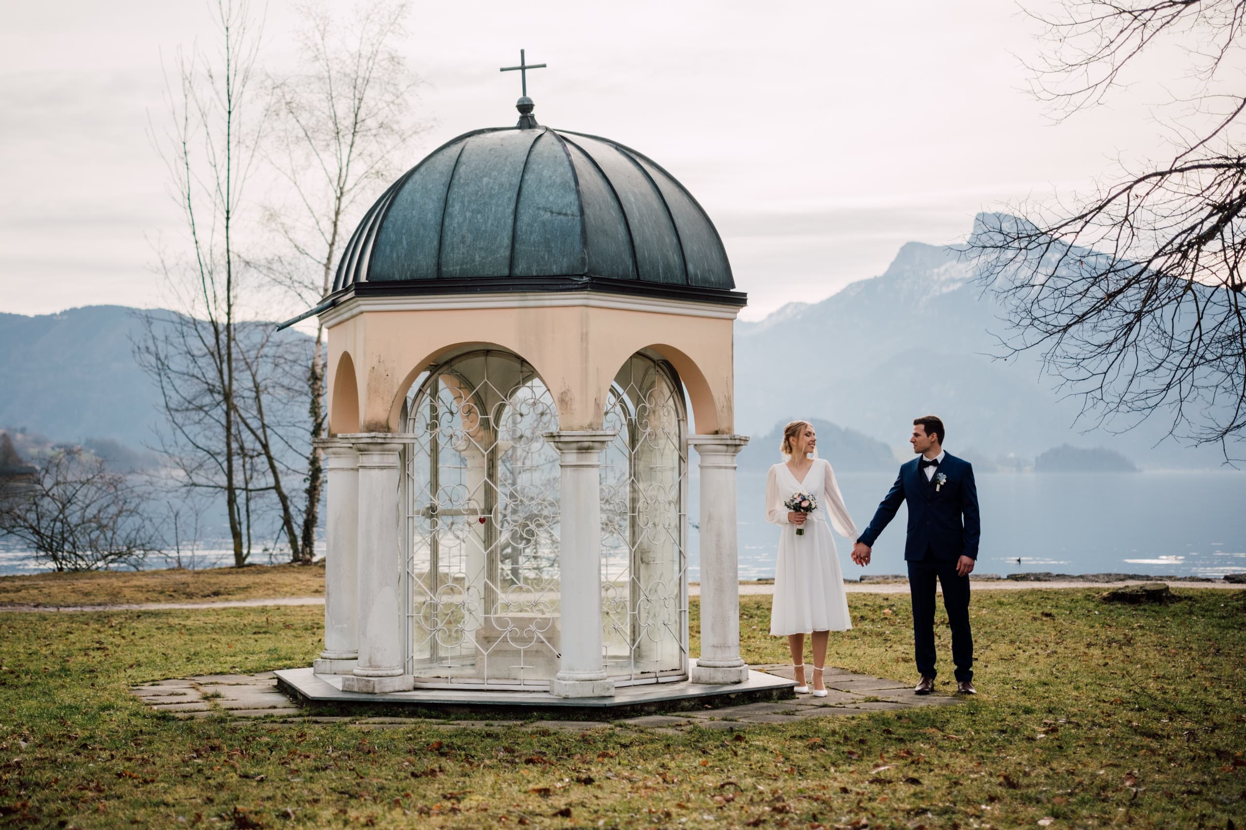 Bride and groom hold hands beside a domed lakeside pavilion with mountains behind